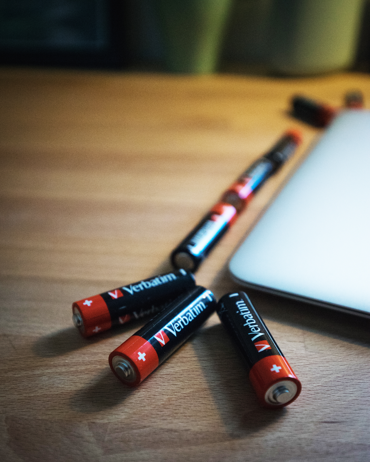 Vier Verbatim AA-batterijen en een pen op een houten tafel, met een gedeeltelijk zichtbare laptop.