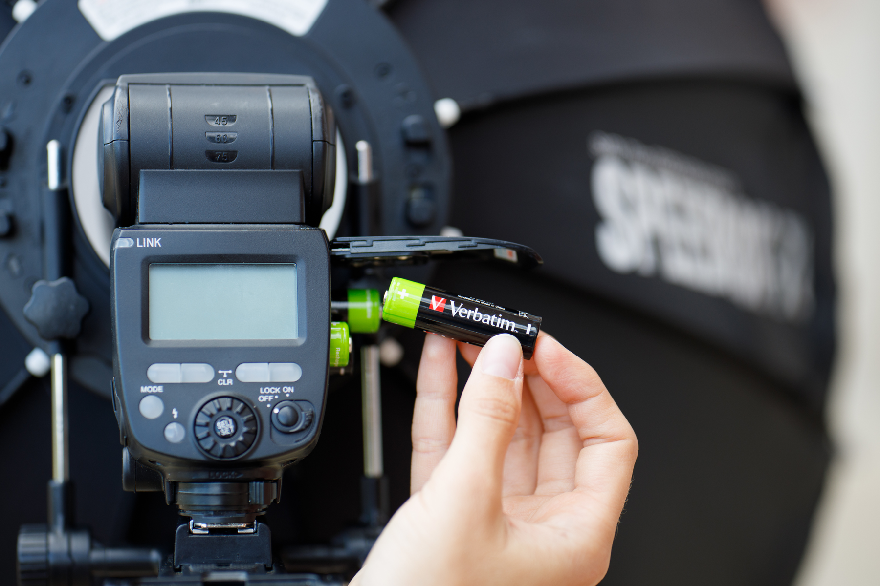 Ręka wkłada baterię AA Verbatim do lampy błyskowej, czarnego urządzenia z ekranem.