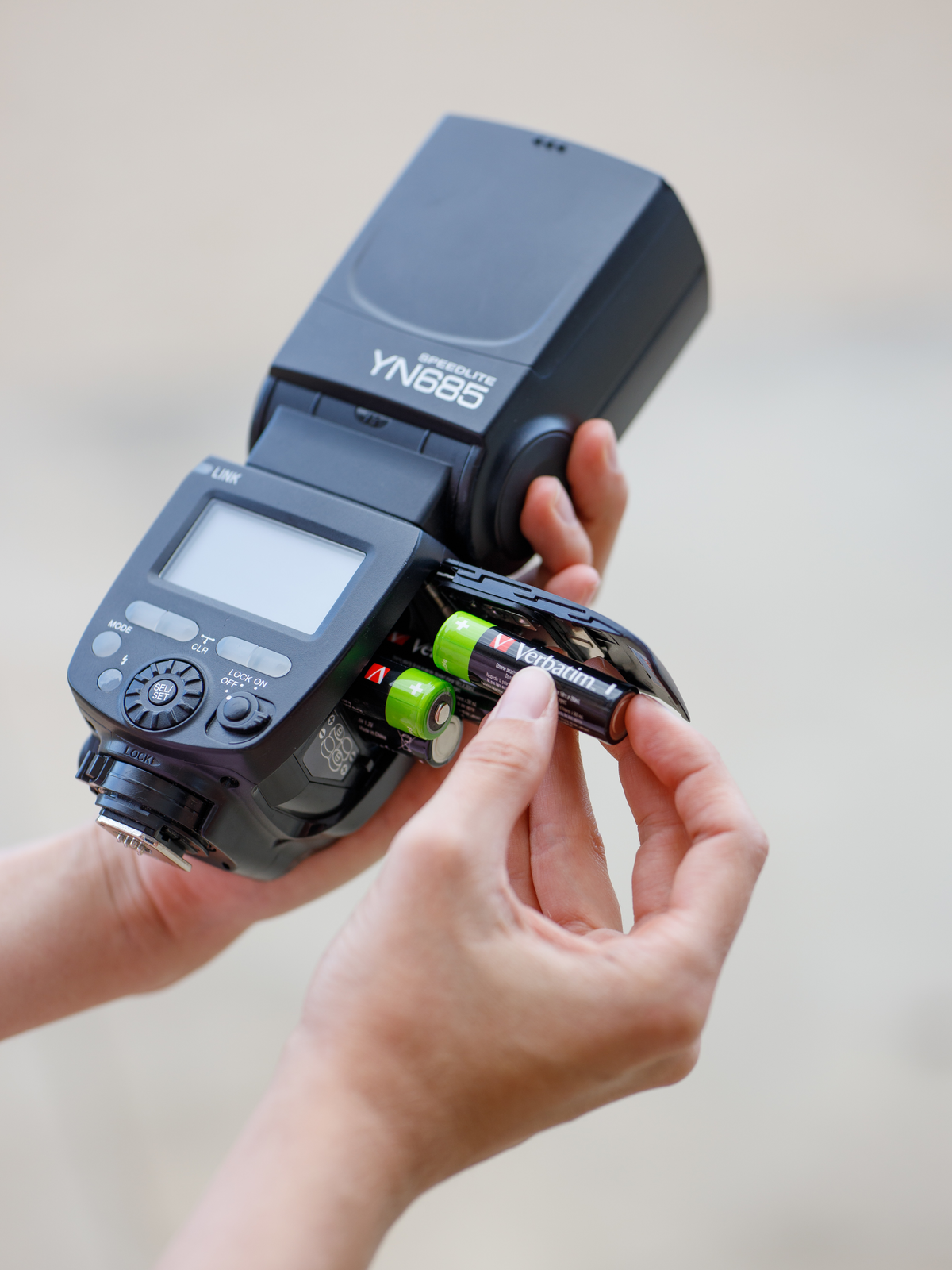 Ręce wkładające baterie AA Verbatim do lampy błyskowej, czarnego urządzenia, na jasnym tle.