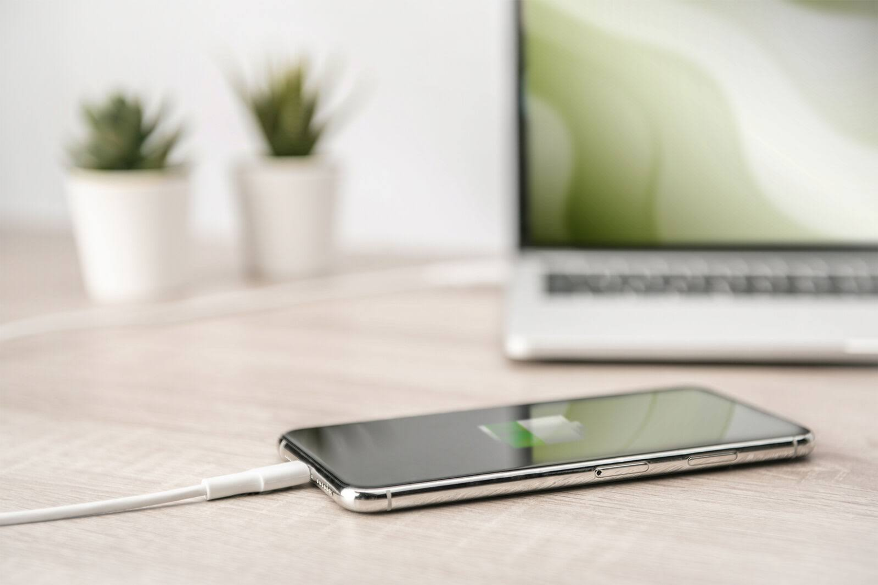 Smartphone en charge sur table en bois, avec un ordinateur portable et des plantes en arrière-plan.