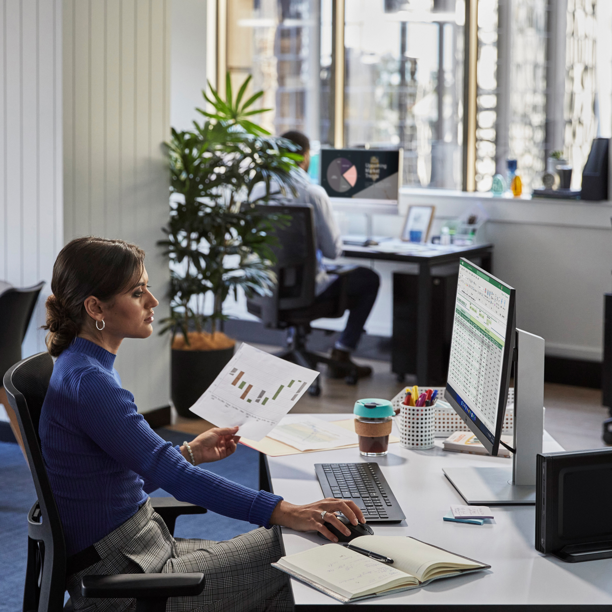 Una donna a una scrivania in ufficio, che lavora al computer. Un'altra persona lavora sullo sfondo.