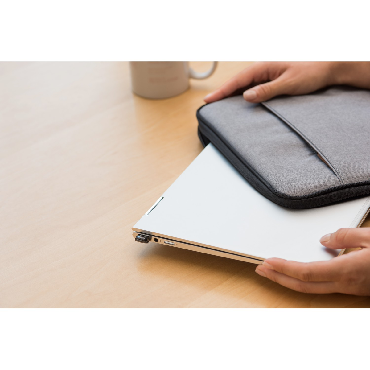 Dłonie trzymające laptopa z szarym etui na jasnej drewnianej powierzchni. Widoczny kubek.
