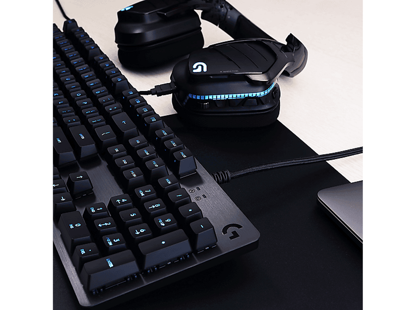 Un teclado, auriculares y ratón negros sobre una superficie negra, con teclas y logos azules brillantes.