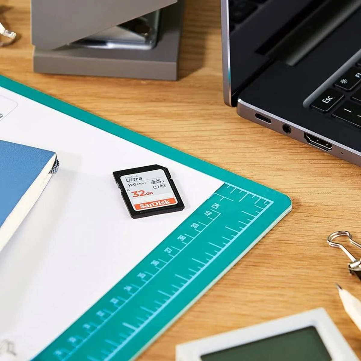 Karta SD na zielonej macie do cięcia. Laptop, zszywacz i notatnik również obecne.