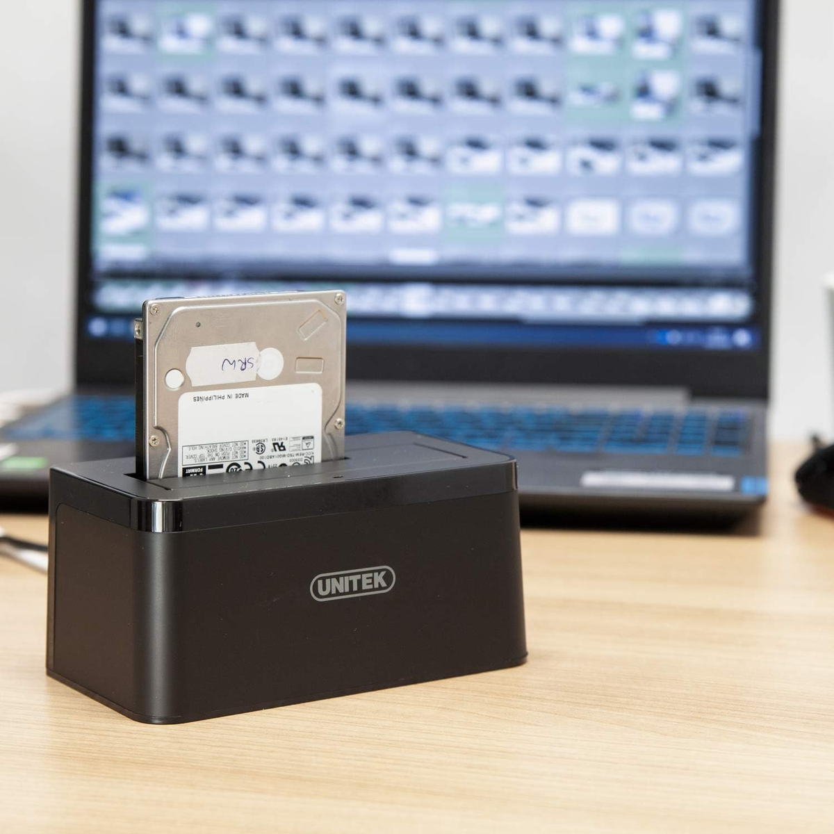 Eine Festplatte wird in ein schwarzes Dock eingesetzt. Ein Laptop mit Bildern ist im Hintergrund.