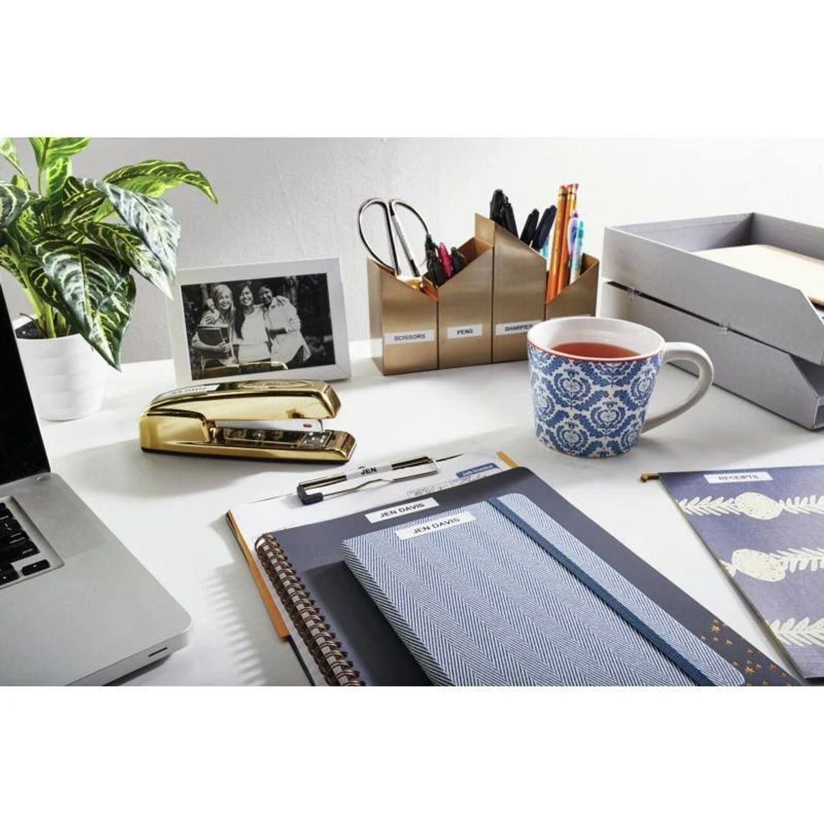 Ein Bürotisch mit verschiedenen Gegenständen: ein Tacker, Stifte, eine Tasse mit Tee, ein Fotorahmen und ein Laptop.