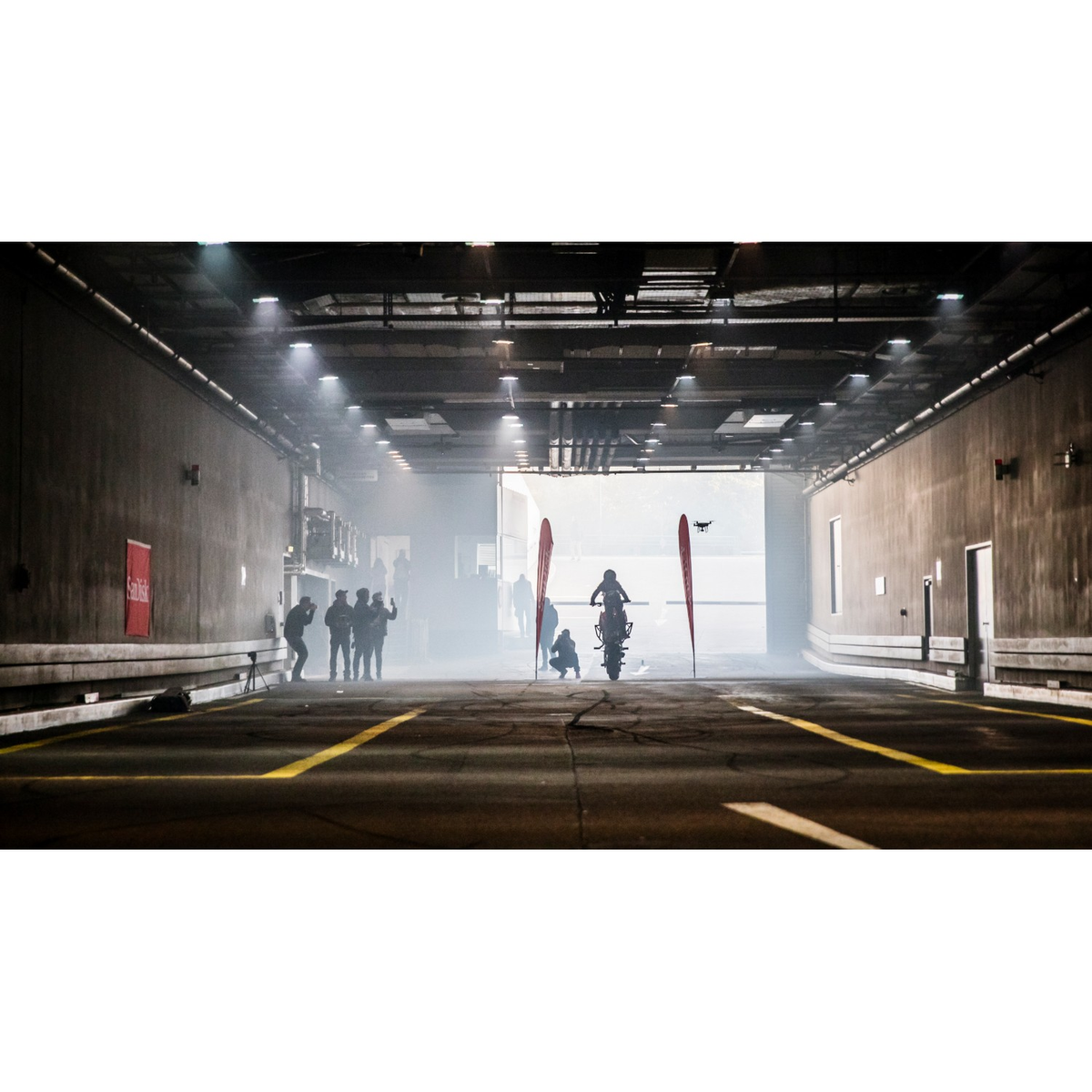 Scena motocyklowa w tunelu z ludźmi. Dron w powietrzu, czerwone flagi.