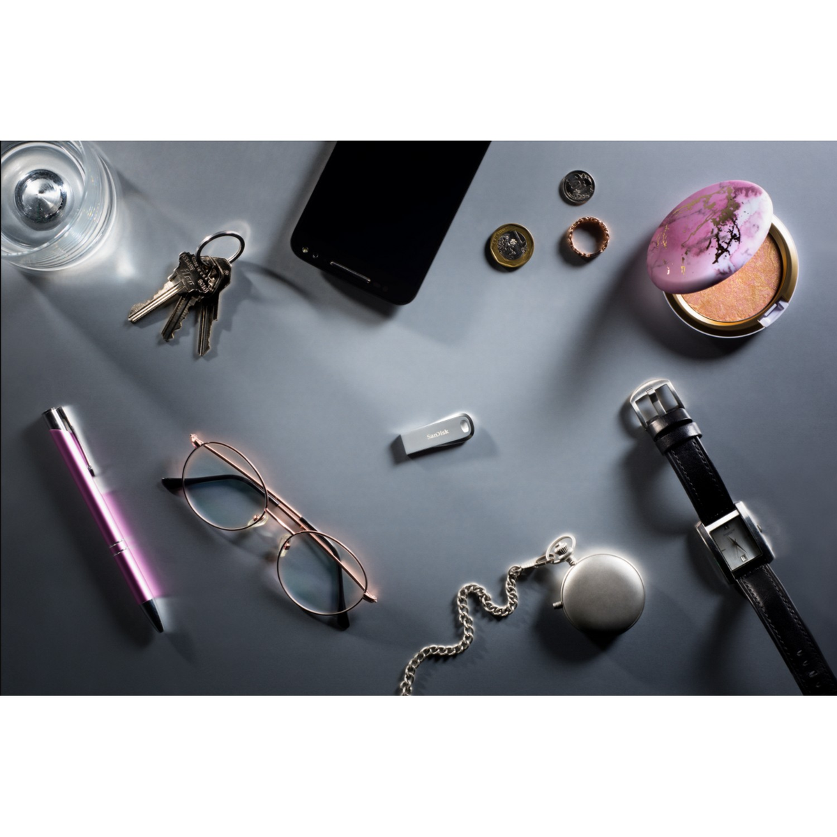 Objets sur une surface grise: clés, lunettes, stylo, montre, pièces, maquillage et clé USB.