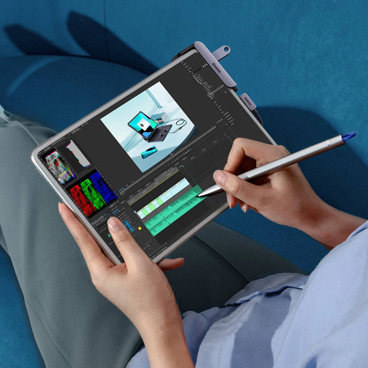 Düzenleme yazılımı açık bir tablet tutan kişi. Ekranda bir kalem kullanılıyor. Arka planda mavi bir kanepe.