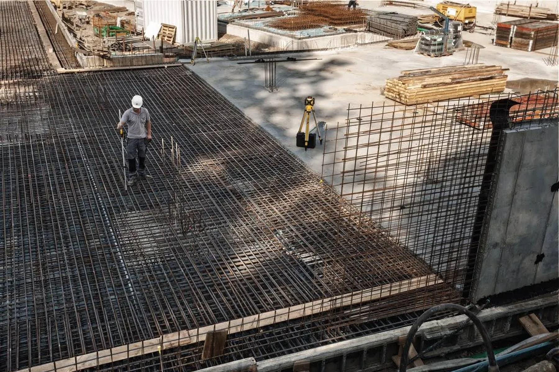 Cantiere edile con operaio, griglia di tondini e attrezzature topografiche.