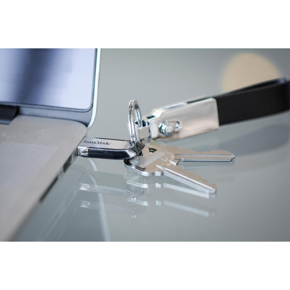 Unidad USB SanDisk y llaves en una superficie de vidrio junto a un portátil.