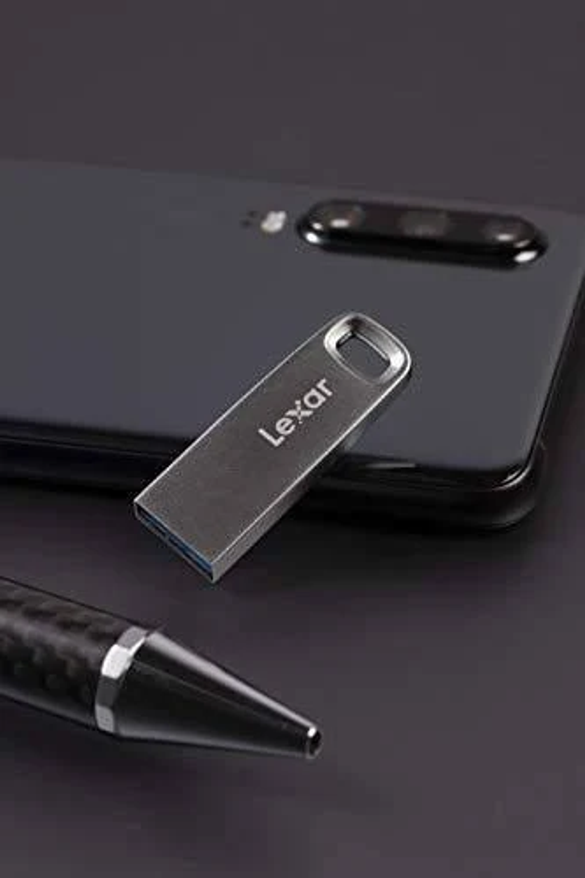 Srebrny pendrive Lexar obok czarnego smartfona i długopisu na ciemnej powierzchni.