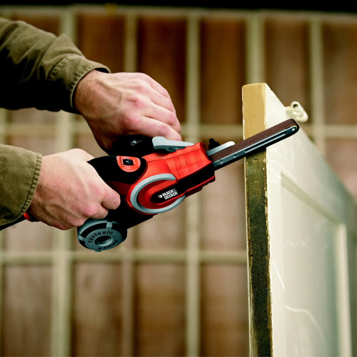 Osoba szlifująca drewnianą ościeżnicę za pomocą czarno-czerwonej szlifierki.
