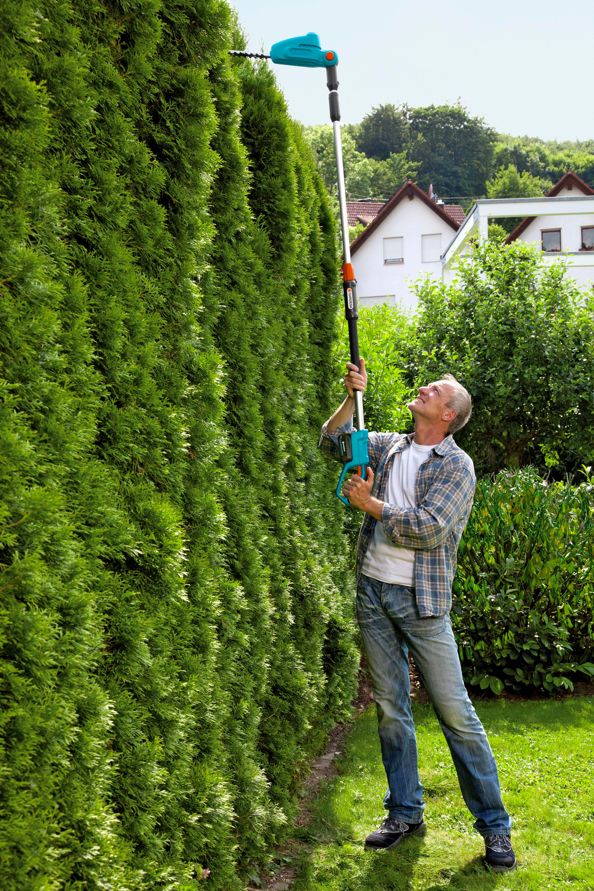 Ein Mann schneidet mit einer elektrischen Heckenschere eine hohe Hecke, in einem Garten.