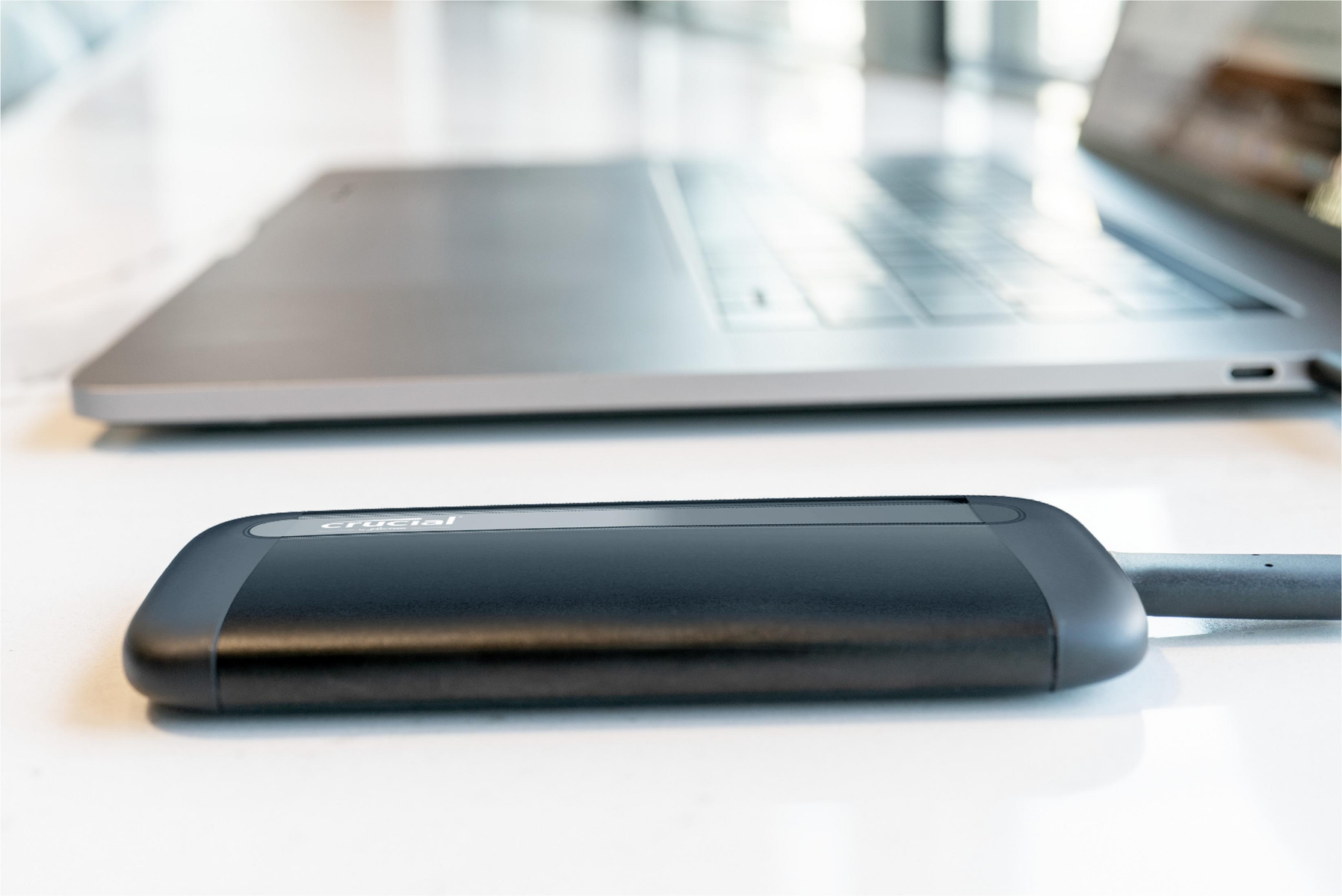 Czarny zewnętrzny dysk twardy Crucial z szarym kablem. W tle laptop.