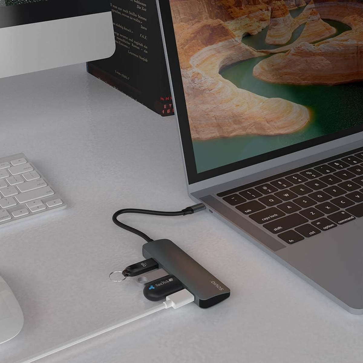 Ein Laptop, der mit einem USB-Hub verbunden ist, mit einem Monitor und einer Tastatur im Hintergrund.