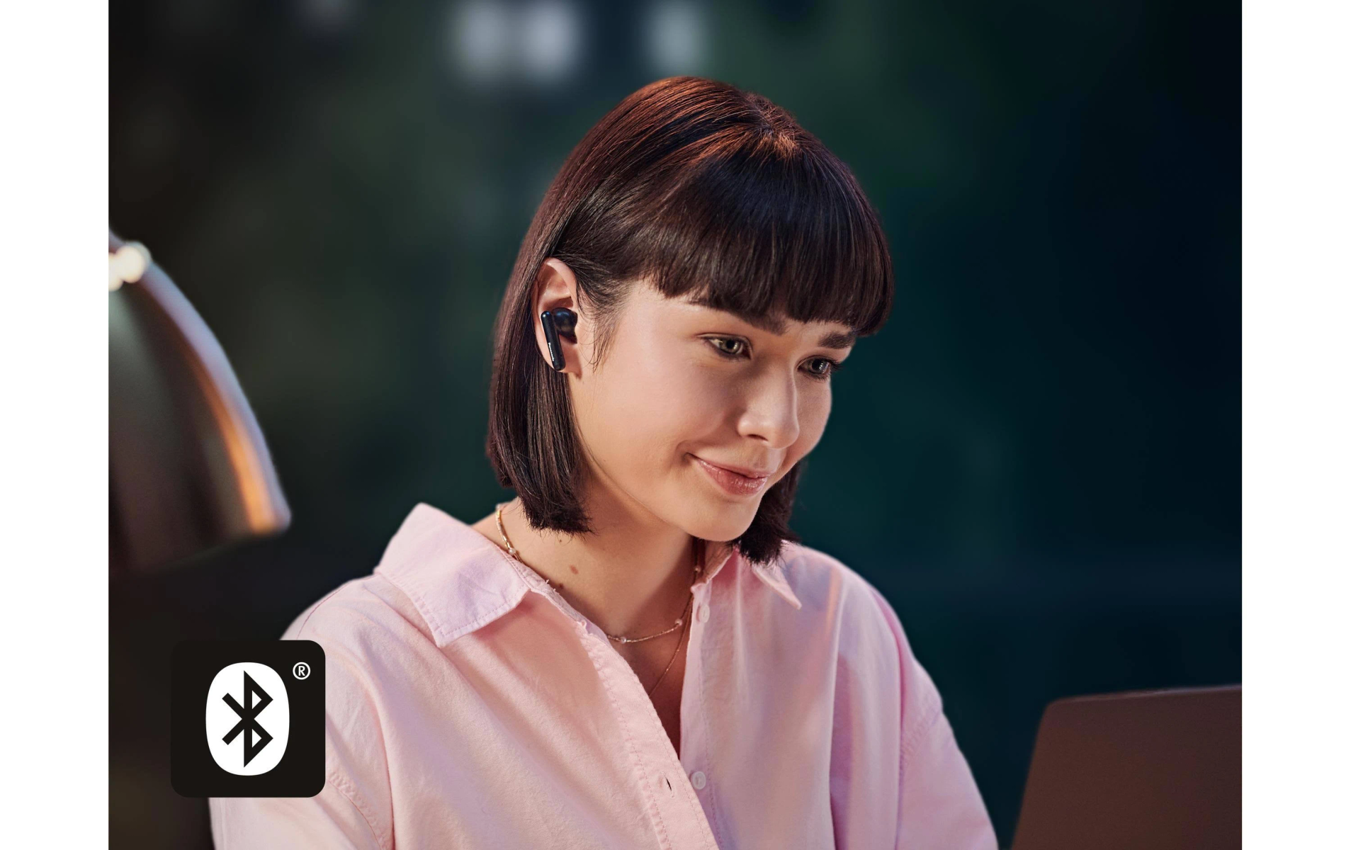Eine Frau mit Ohrhörern lächelt einen Laptop an. Ein Bluetooth-Symbol ist in der Ecke. Sie trägt ein rosa Hemd und eine Halskette.
