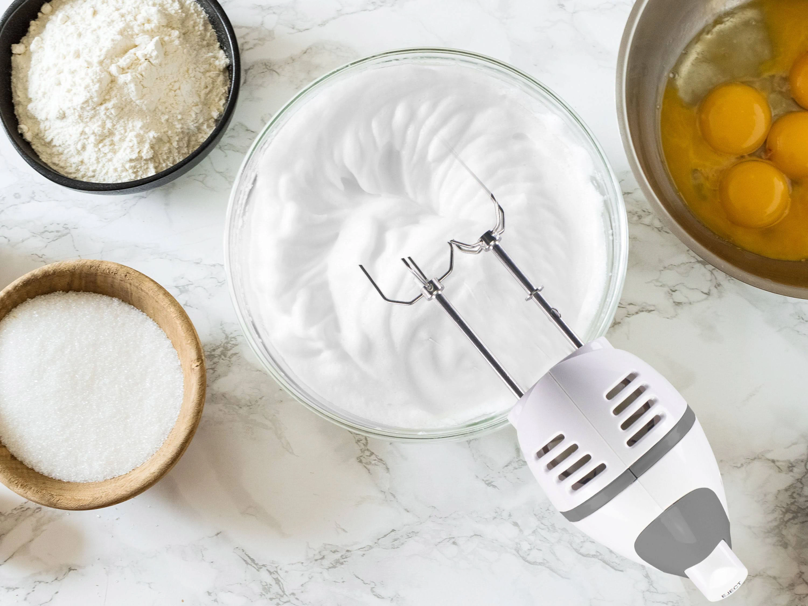 Ingredienti per cuocere: farina, zucchero, uova e albumi montati. Un mixer è al centro.