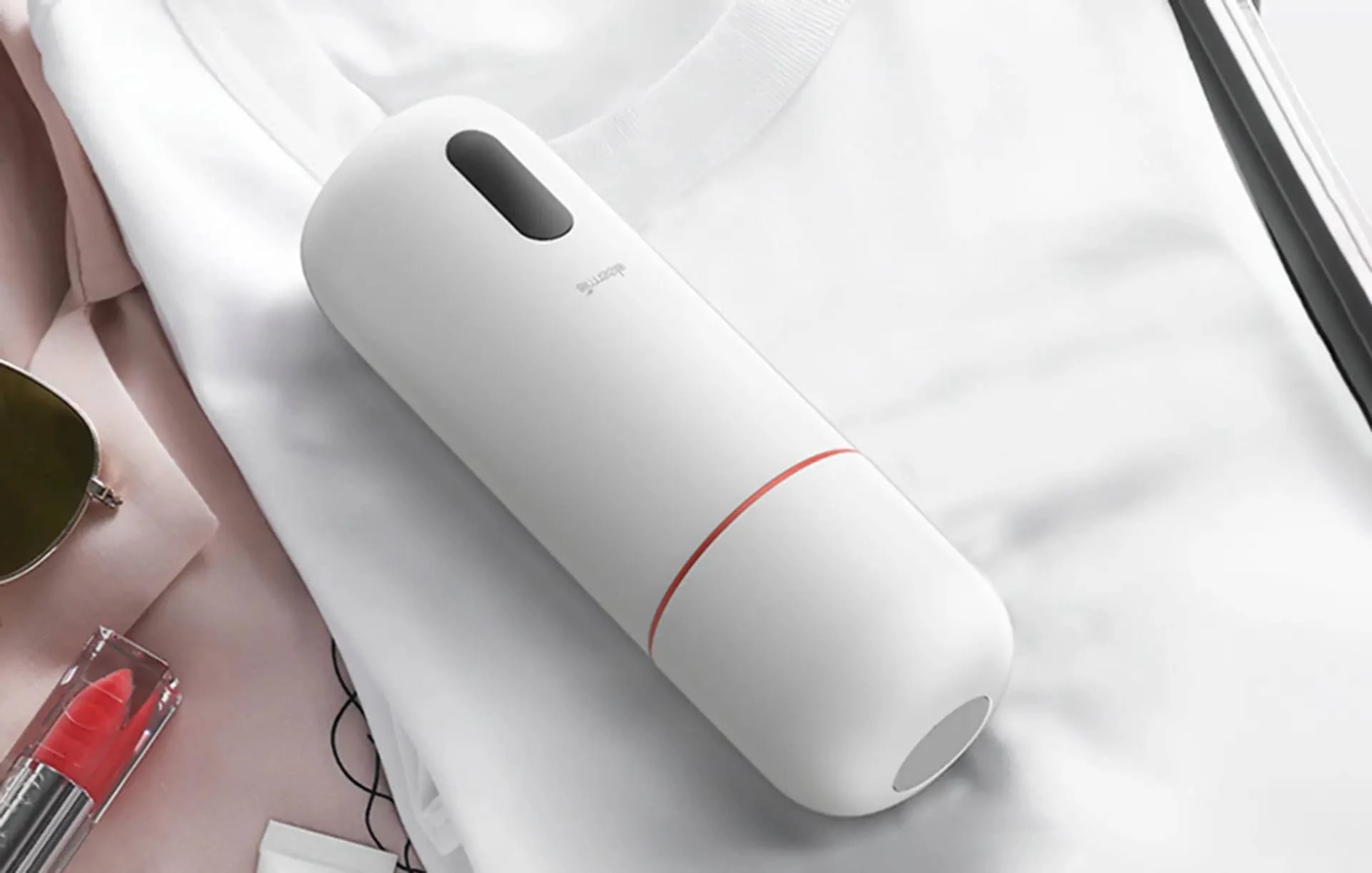 Biała przenośna parownica do ubrań na białej koszuli z szminką i okularami przeciwsłonecznymi w tle.