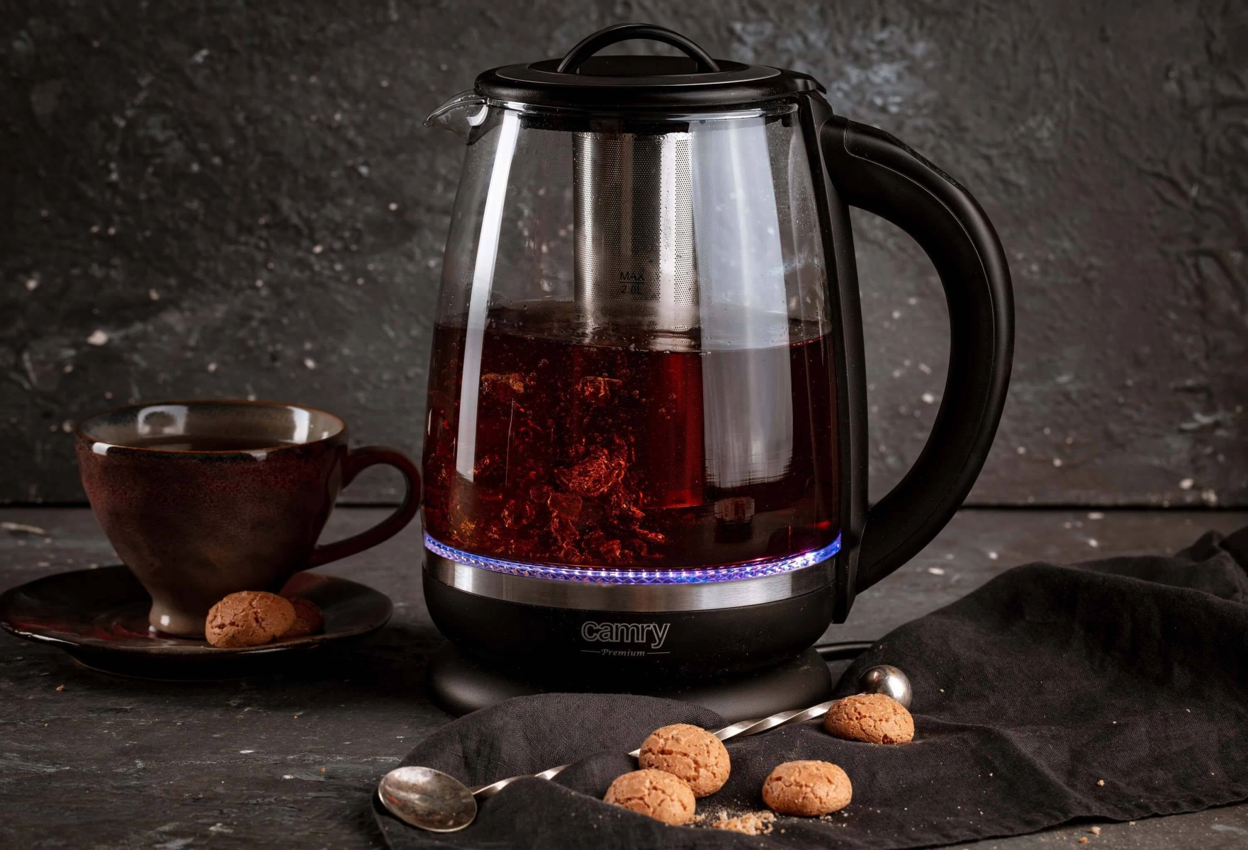 Szklany czajnik elektryczny z herbatą w środku i kubkiem. Ciasteczka i łyżka. Ciemne tło.