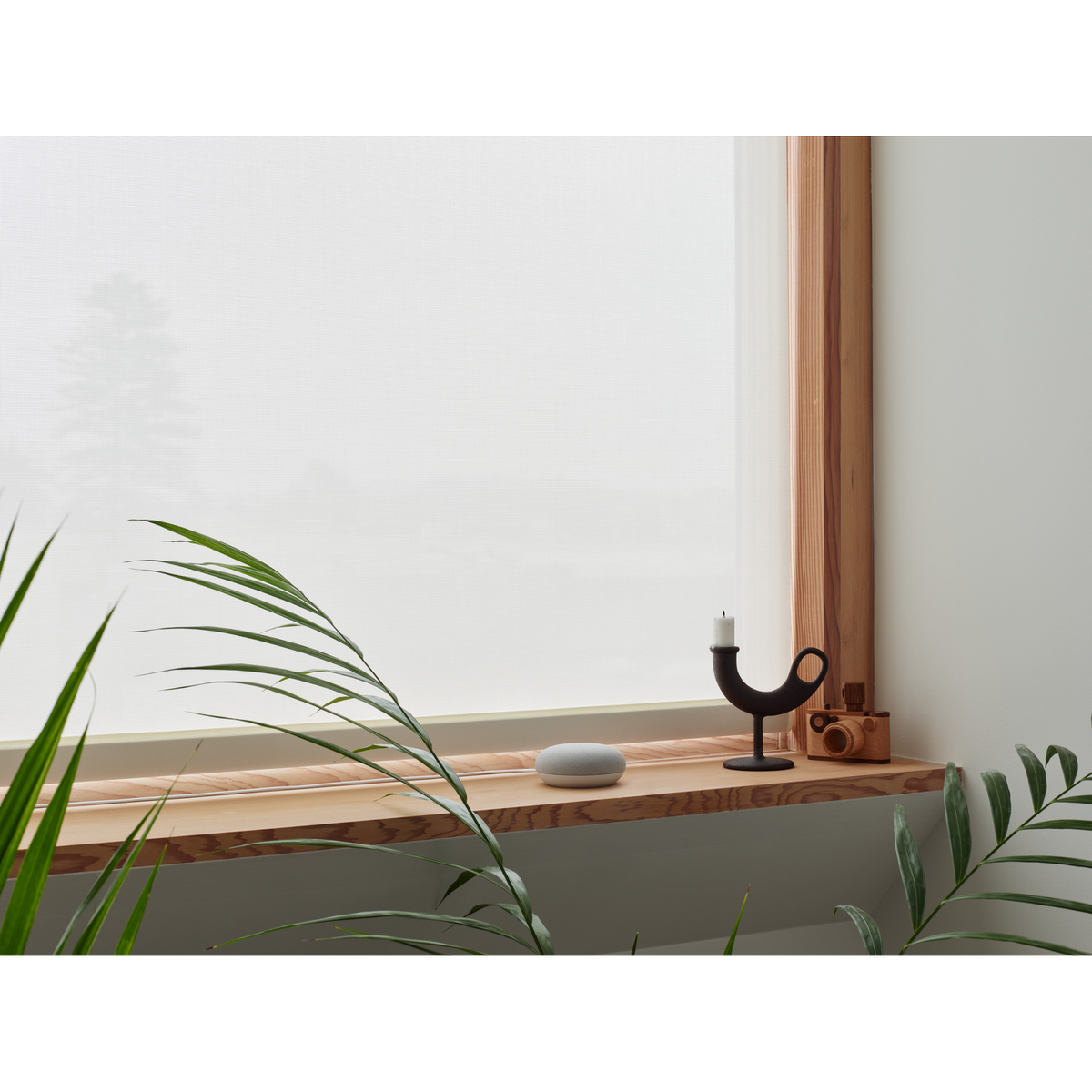 Okno z białą roletą, świecą i drewnianym aparatem. Roślina i głośnik inteligentny na półce.