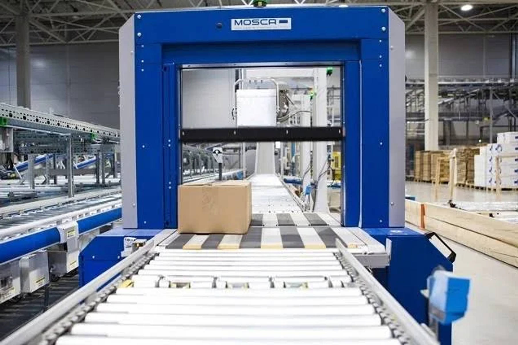 Große blaue Industriemaschine mit Förderbändern. Ein Karton steht in der Mitte des Förderbands.