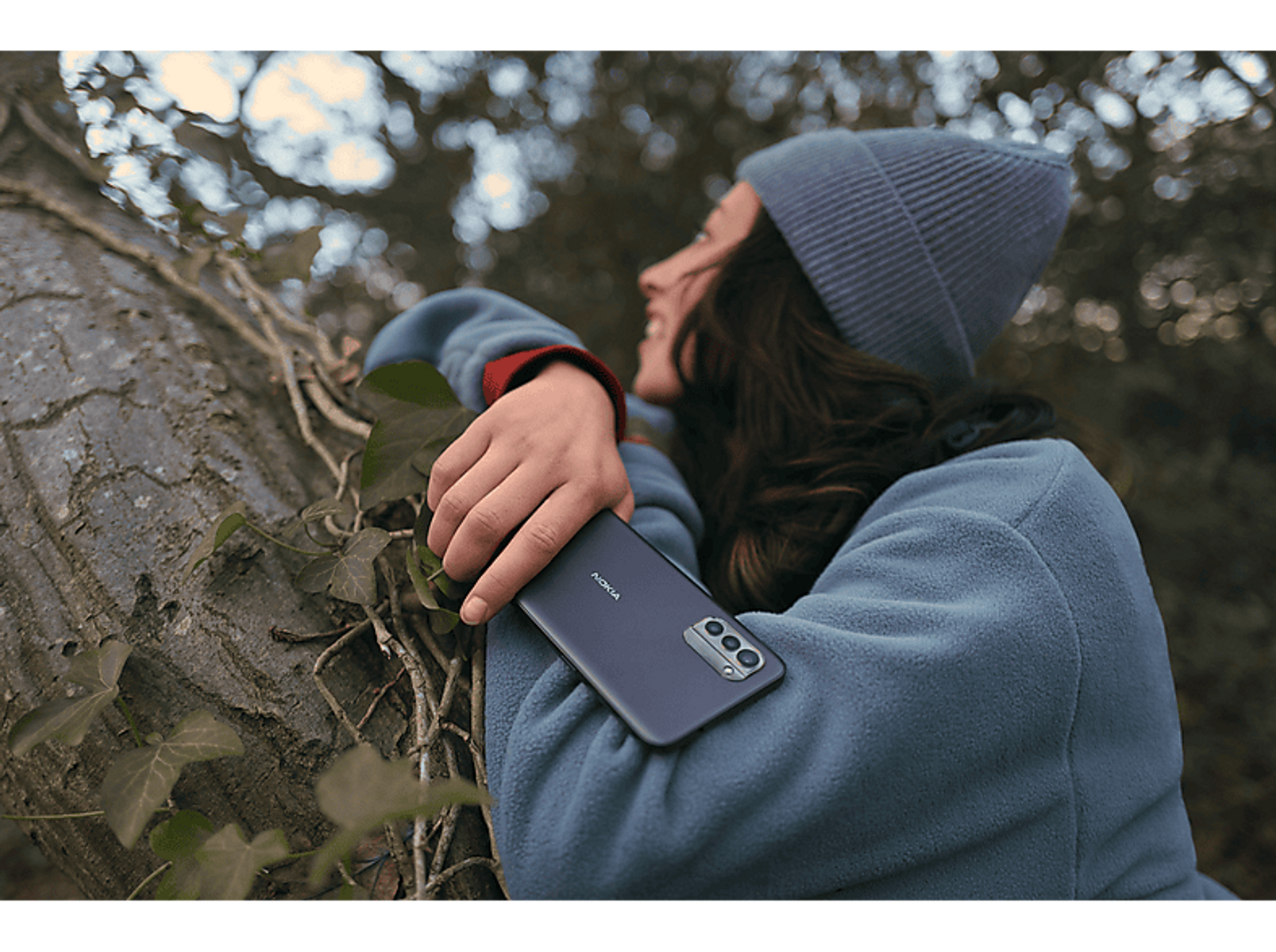 Kobieta z telefonem oparta o drzewo. Niebieska czapka i kurtka. Na zewnątrz.