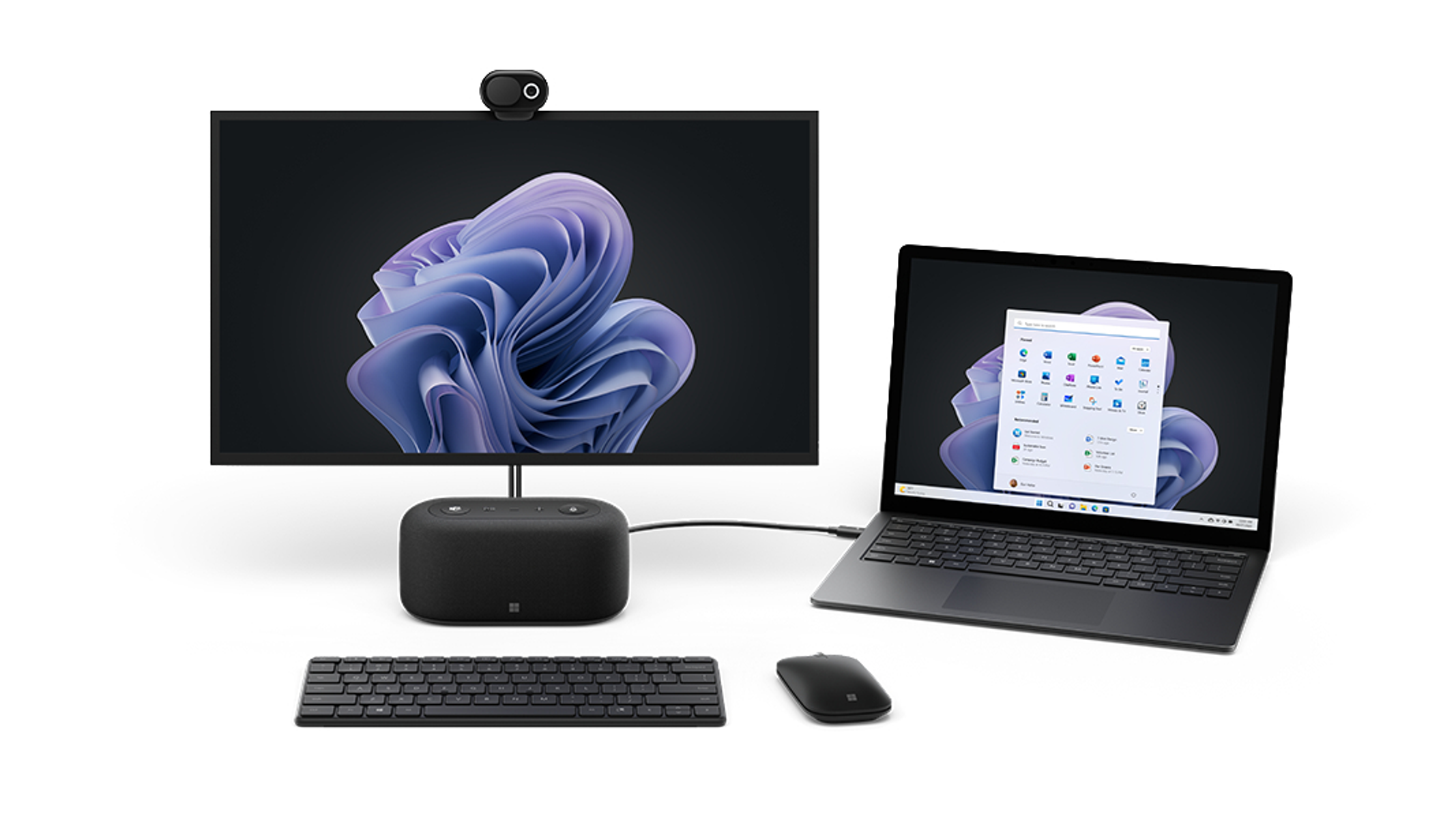 Configurazione computer Microsoft con monitor, laptop, altoparlante, tastiera e mouse su una superficie bianca.