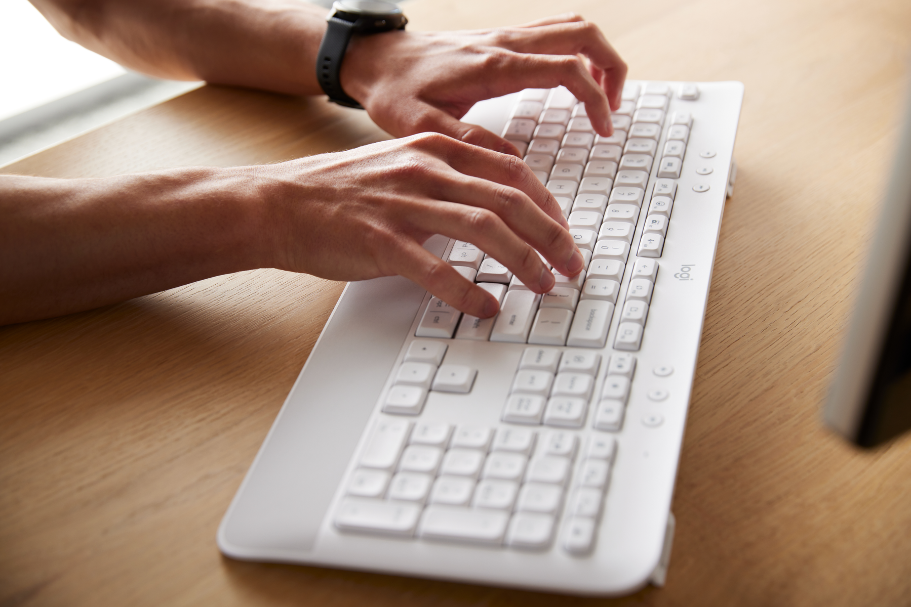 Dłonie piszące na białej klawiaturze. Zegarek na lewym nadgarstku. Tło drewniane.