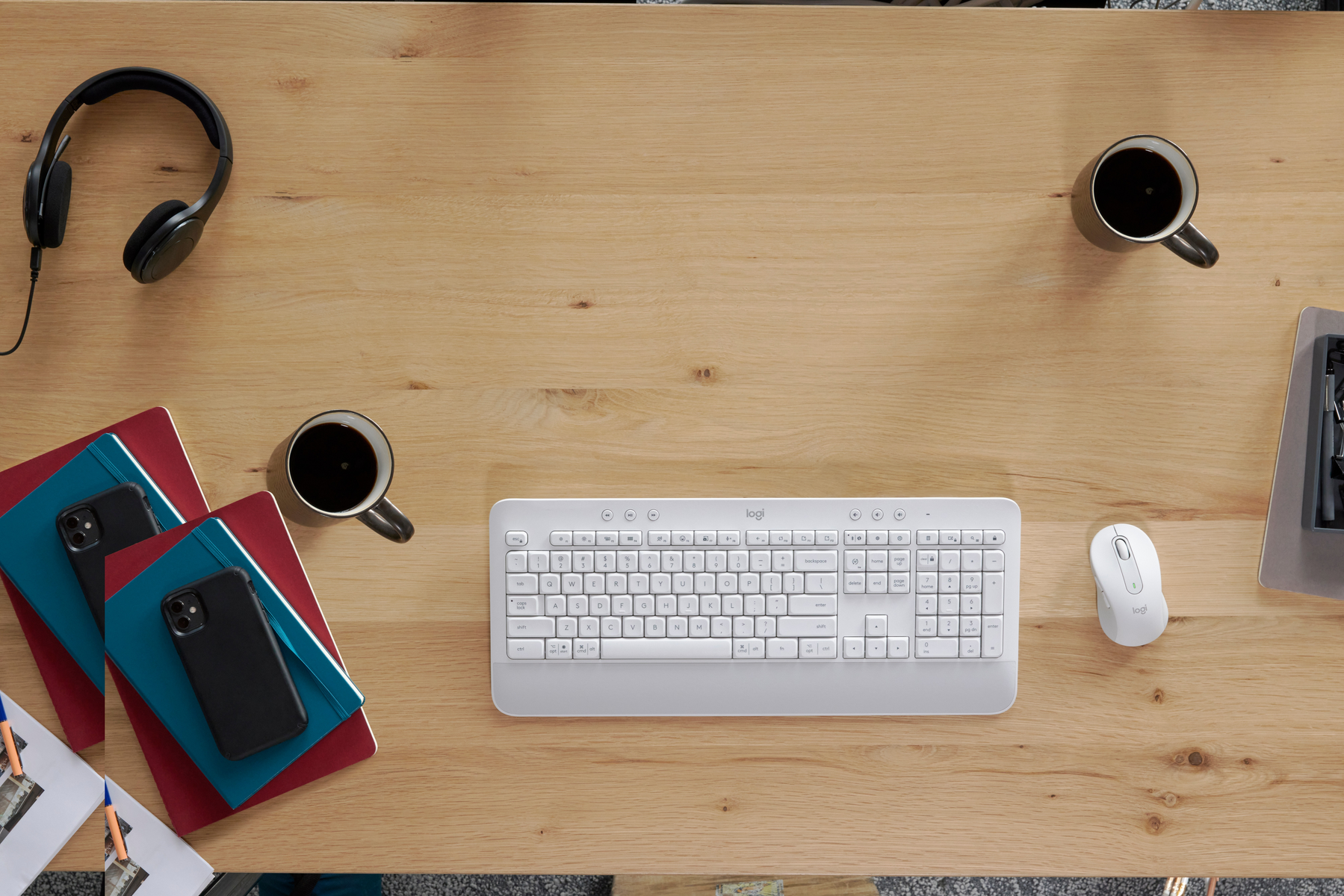 Widok z góry na biurko z klawiaturą, myszką, telefonem, notatnikami i kawą.