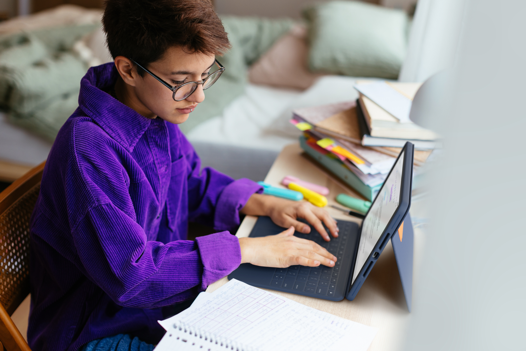 Junge Person, die an einem Laptop arbeitet. Trägt eine Brille und ein lila Hemd. Bücher und Papiere auf dem Schreibtisch.