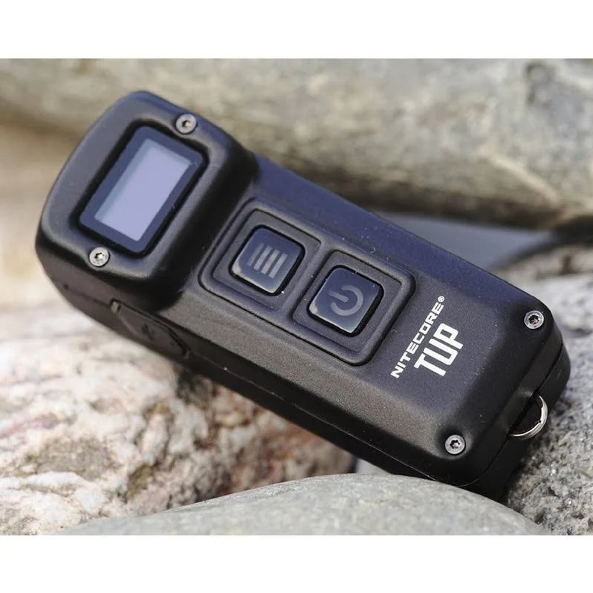 Schwarze Nitecore TUP Taschenlampe auf Felsen. Zwei Knöpfe, Display und Schlüsselringschlaufe.
