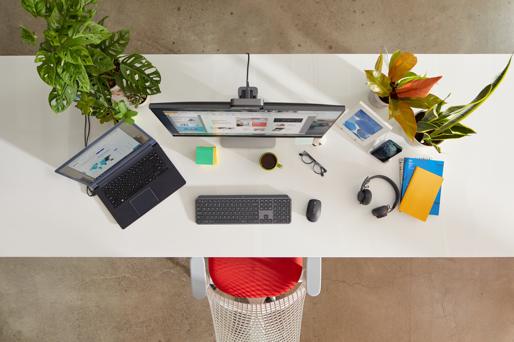 Widok z góry na biurko z laptopem, monitorem, roślinami i artykułami biurowymi, na białym tle.