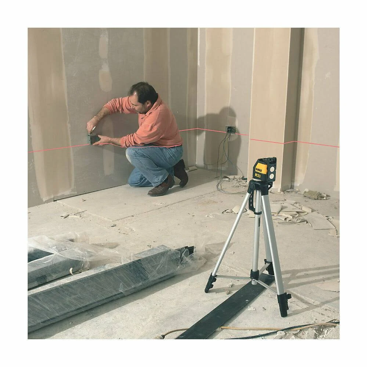 Diz çökmüş adam, lazer terazisi kullanıyor. Kırmızı lazer çizgisi duvarda. Tripod ve enkaz önde.