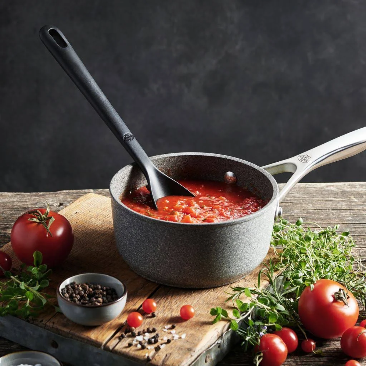 Ein Topf mit Tomatensauce mit einem Löffel, umgeben von Tomaten, Kräutern und Gewürzen auf einem Holzbrett.