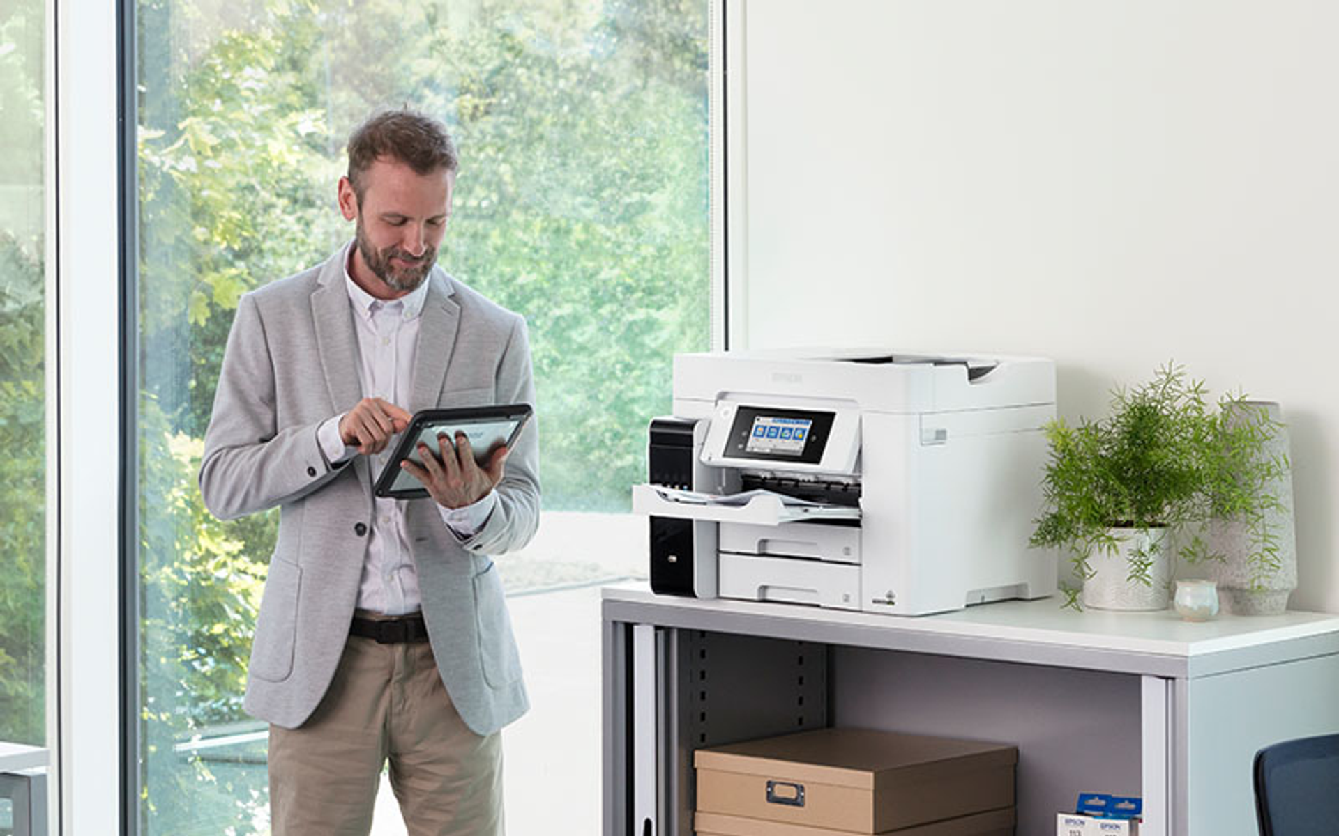 Mężczyzna używa tabletu w pobliżu drukarki. Otoczenie biurowe z roślinami i oknem.