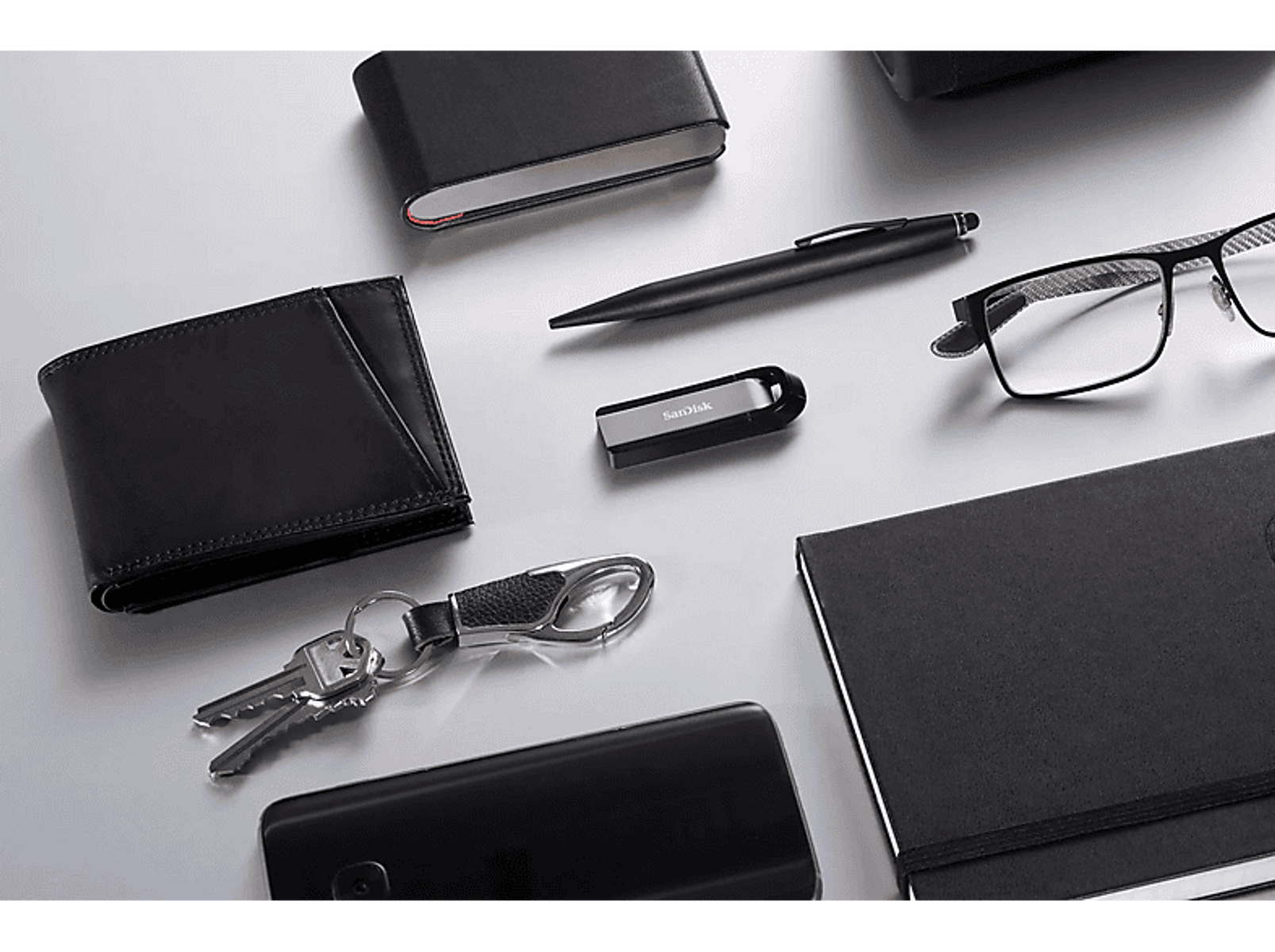 Czarne akcesoria: portfel, długopis, pendrive, okulary, klucze, telefon i notatnik na białym tle.