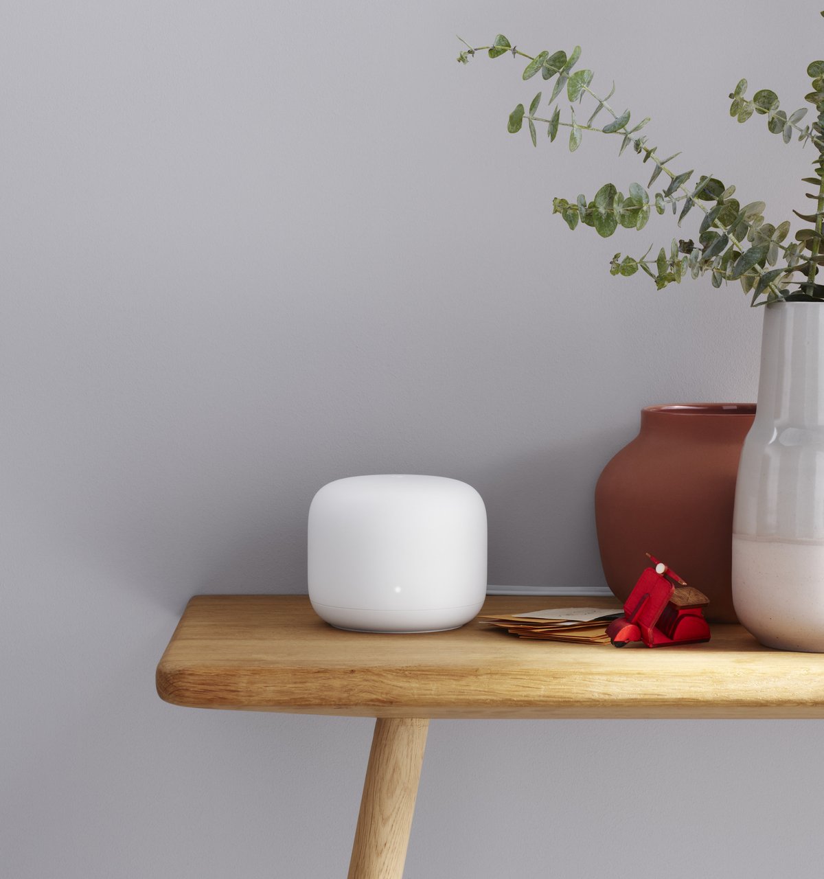 Biały router Google Nest na drewnianym stoliku z rośliną i czerwonym skuterem obok.