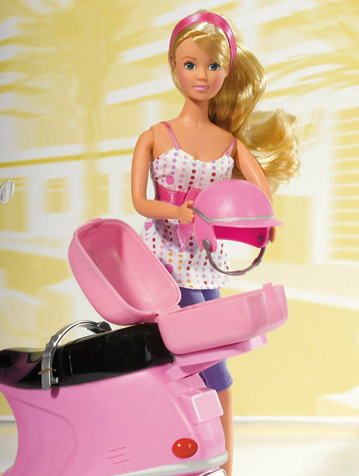 Blonde Puppe mit pinkfarbenem Roller und Helm. Trägt ein gepunktetes Kleid.