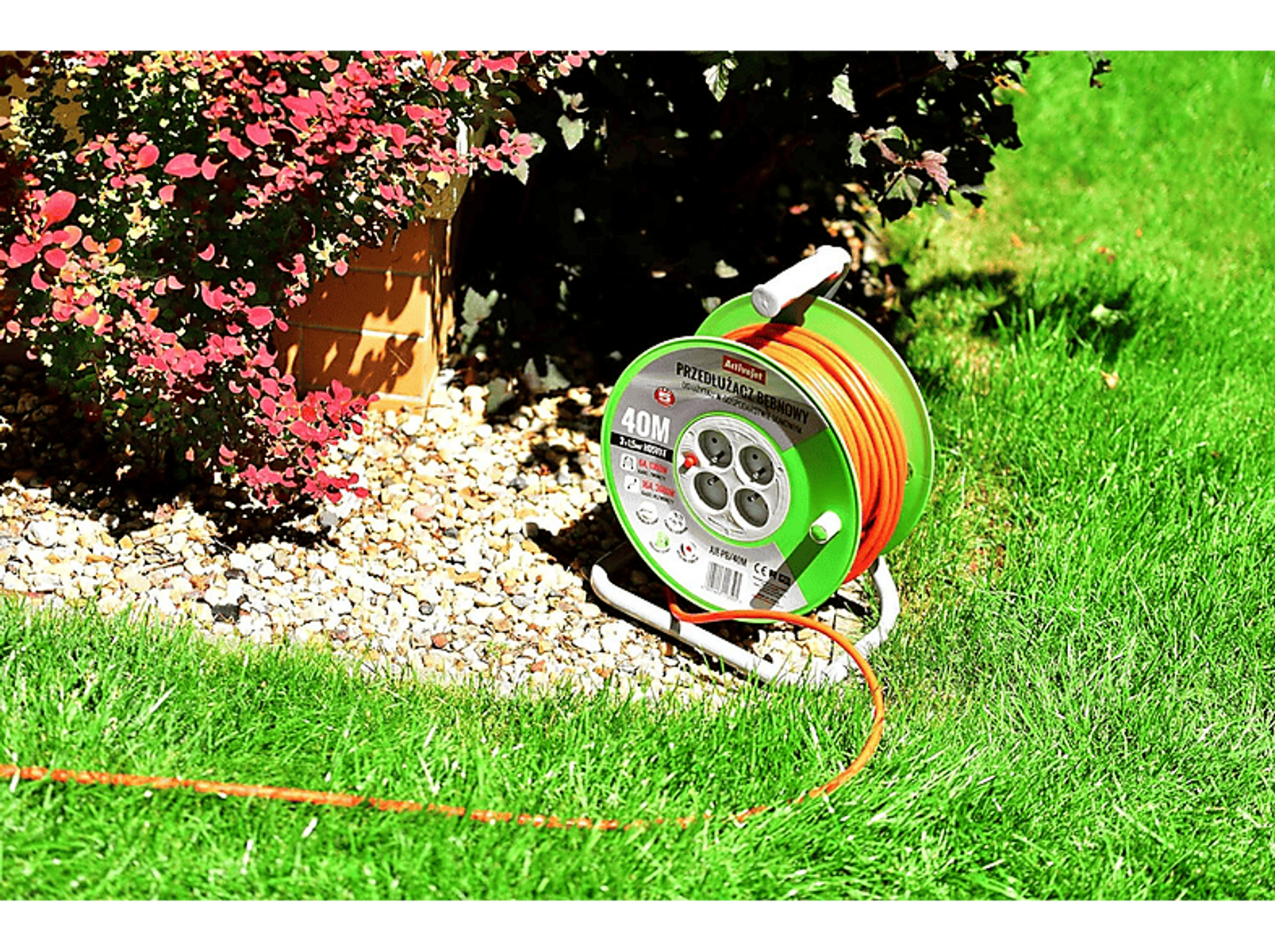 Avvolgicavo verde sull'erba. Cavo arancione avvolto e collegato. Arbusto rosso sullo sfondo.
