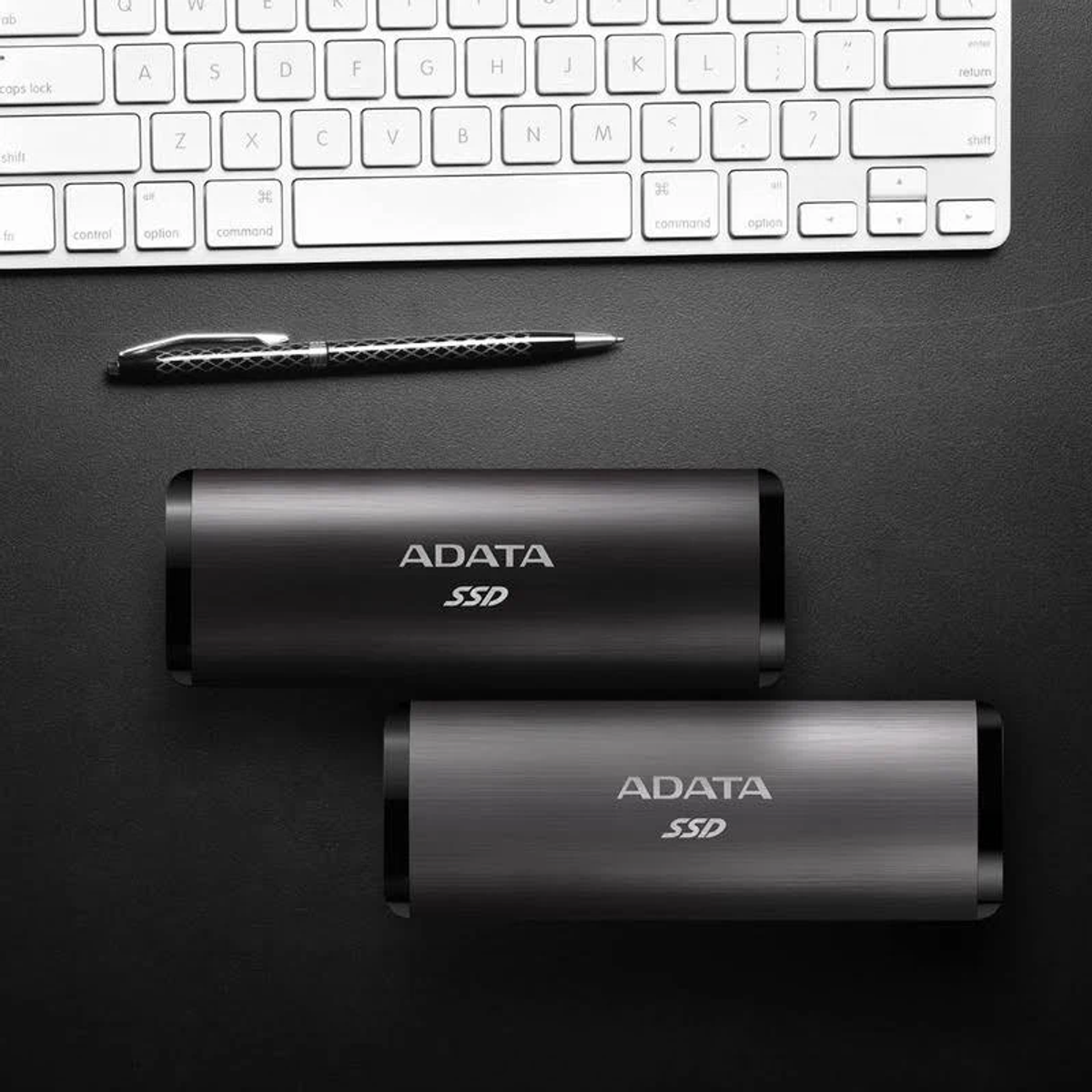 Dwa czarne dyski SSD ADATA na czarnej powierzchni, z długopisem i klawiaturą w tle.