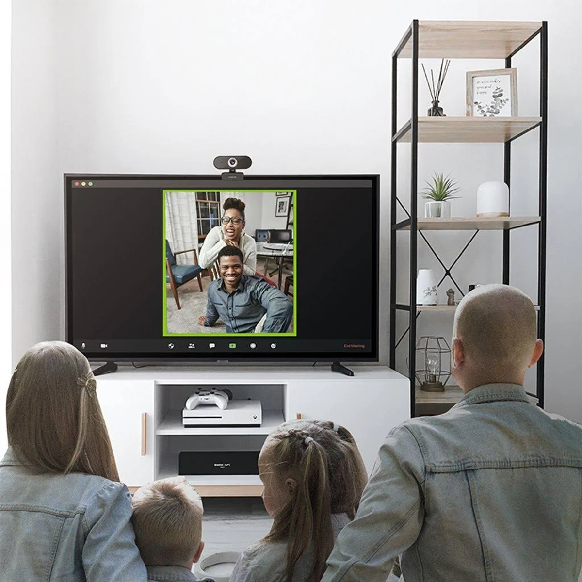 Rodzina oglądająca rozmowę wideo w telewizji. Kamera internetowa znajduje się na górze telewizora. Półka z dekoracjami po prawej stronie.