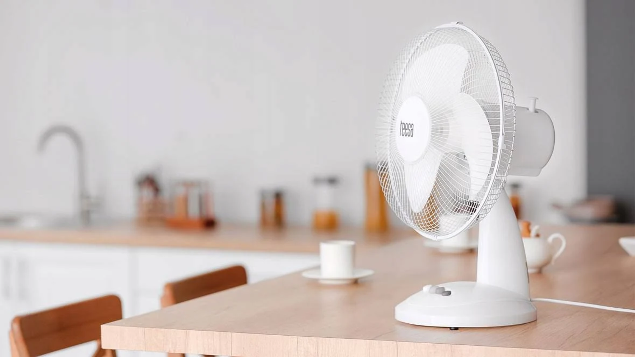 Weißer elektrischer Ventilator auf einem Holztisch in einer hellen Küchenumgebung.