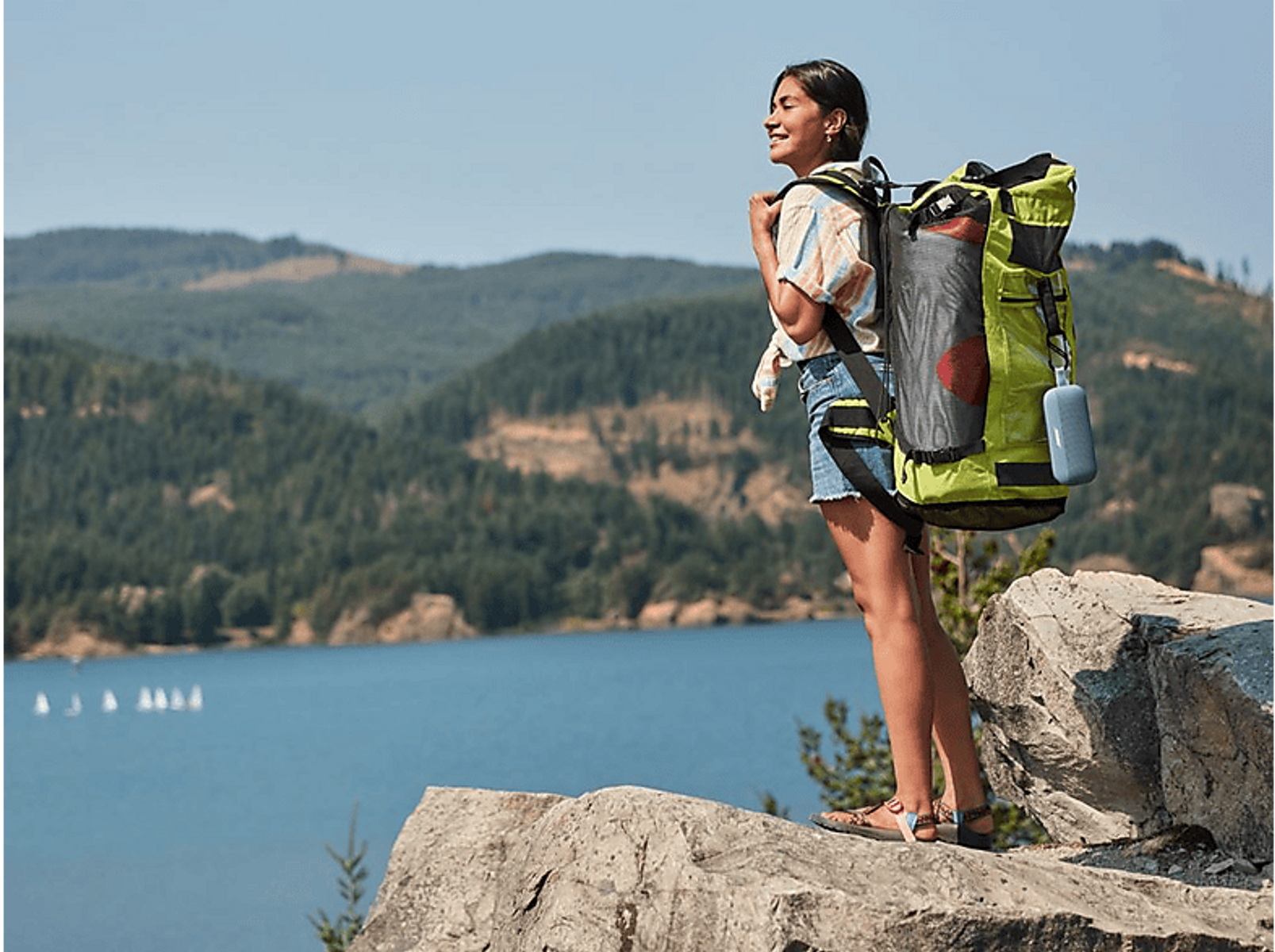 Mujer de senderismo con mochila y altavoz azul en una roca. Montañas y lago al fondo.