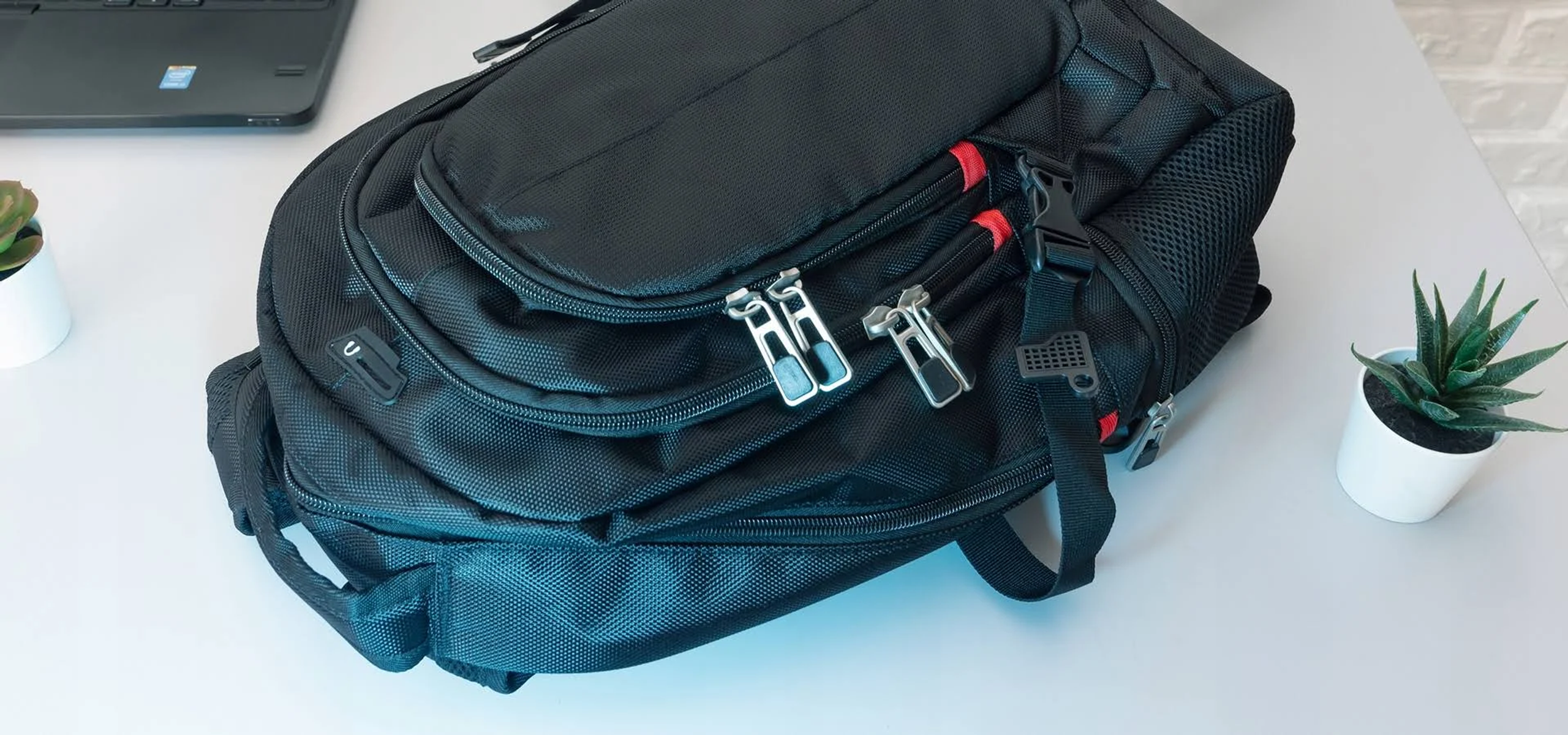 Ein schwarzer Rucksack mit silbernen Reißverschlüssen auf einem weißen Schreibtisch. Links eine kleine Pflanze.