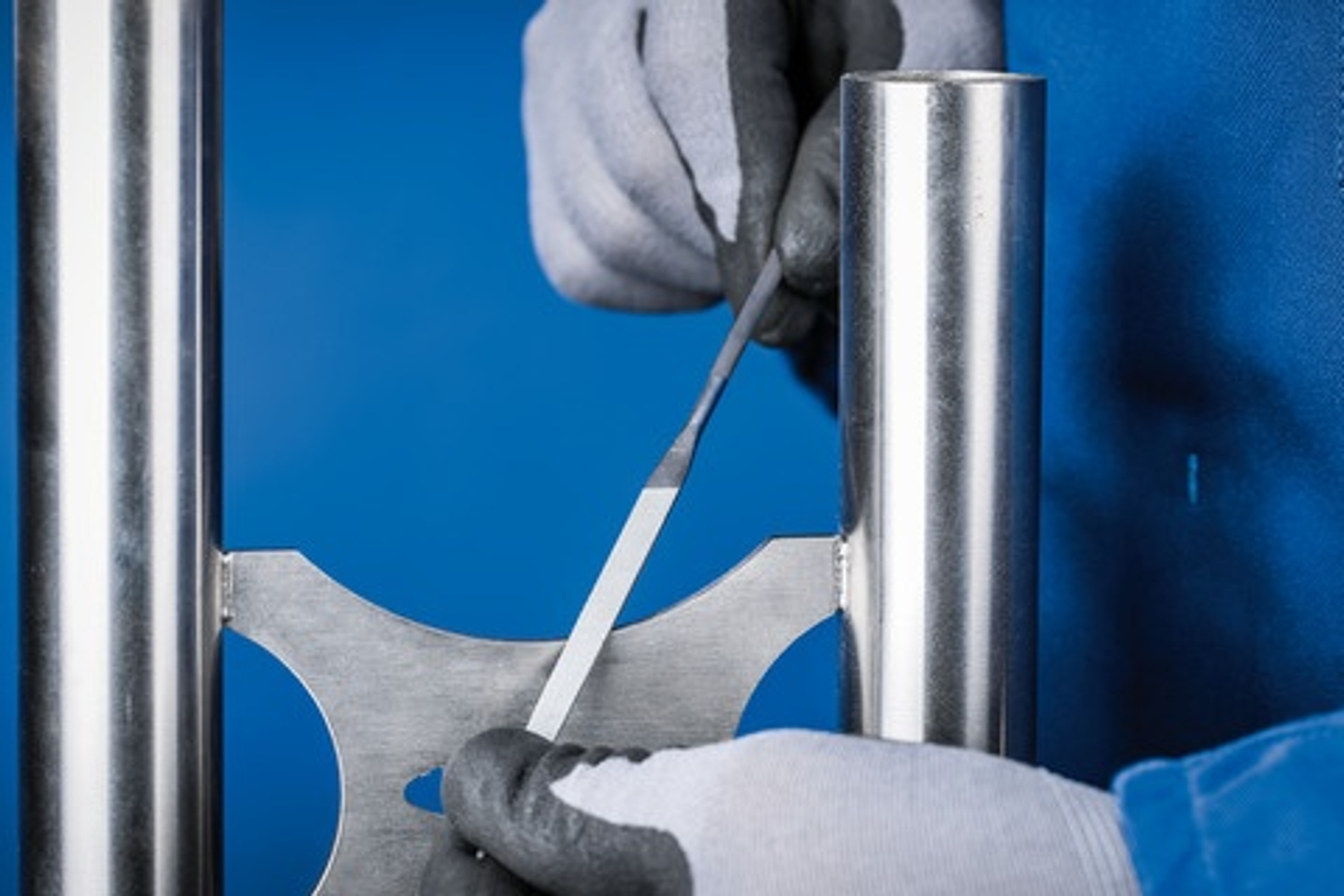 Hände mit Handschuhen, die ein Metallstück mit einer Feile bearbeiten. Die Szene ist in einer Werkstatt.