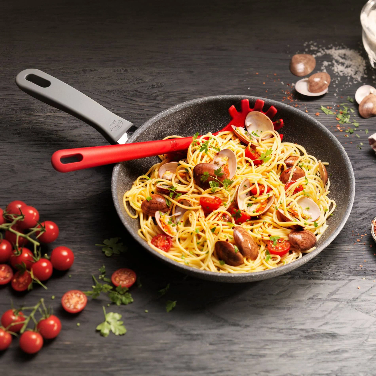 Spaghetti mit Muscheln und Tomaten in einer Pfanne mit rotem Servierlöffel.