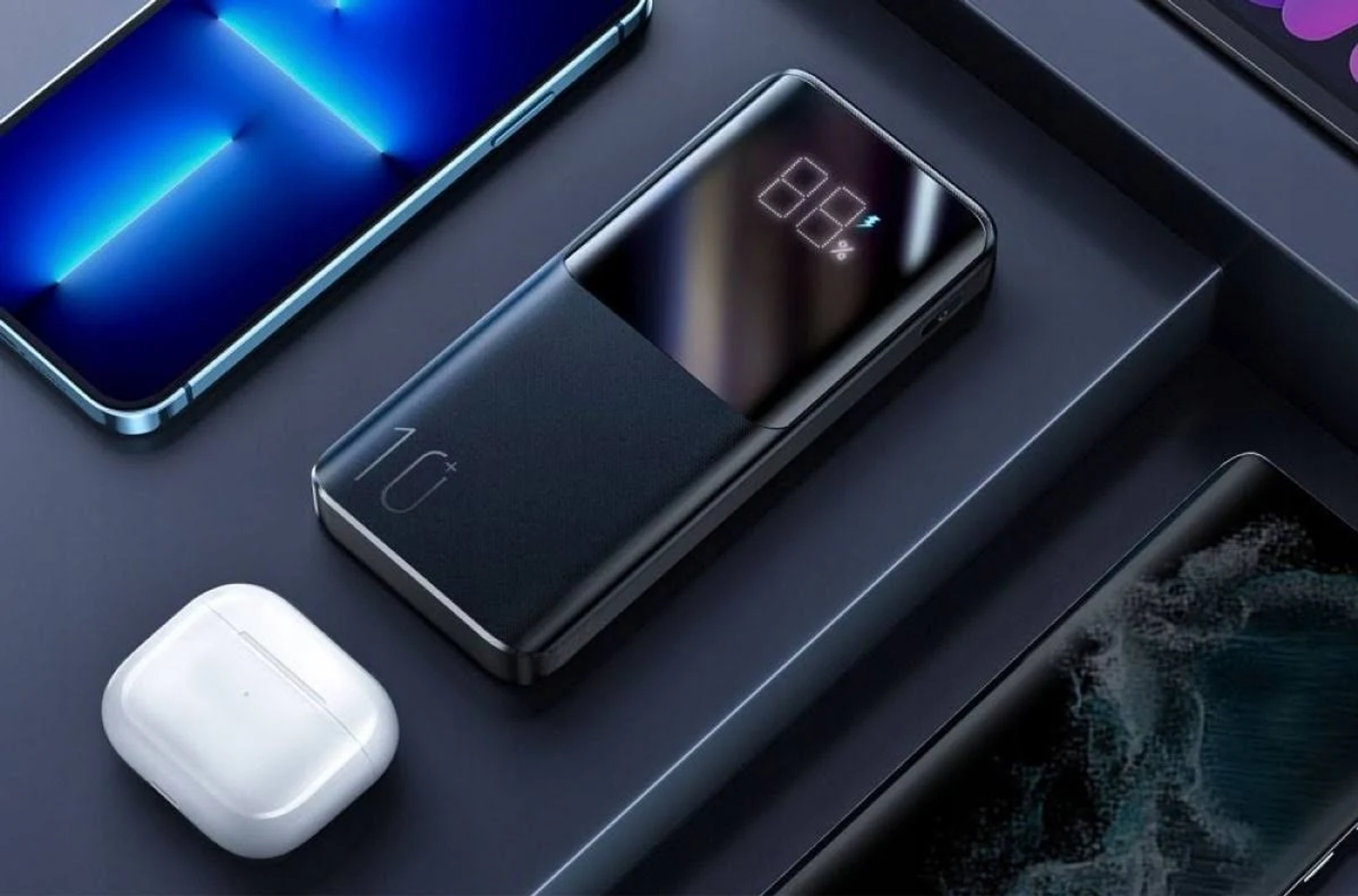 Power bank z wyświetlaczem, telefonem i bezprzewodowymi słuchawkami na ciemnej powierzchni.