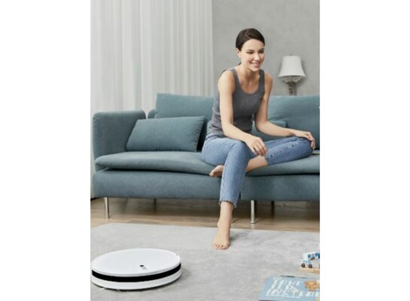 Mujer sentada en sofá mirando robot aspirador. La habitación tiene una alfombra.