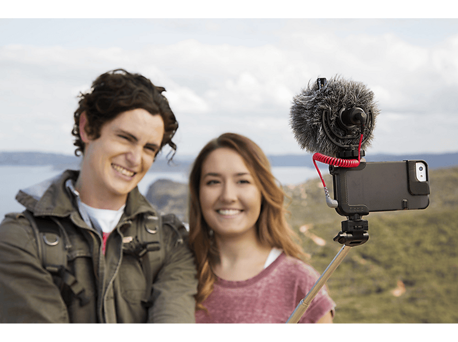 Dos personas sonriendo, sosteniendo un teléfono con un micrófono. Están en una colina con una hermosa vista.